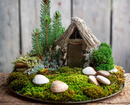 Child's simple fairy garden with rustic house, seashells, moss lawn and mini trees made of fir tree fronds.