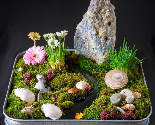 Fairy garden on a shallow tin box base with shells, feature rock, stream, and flowers.