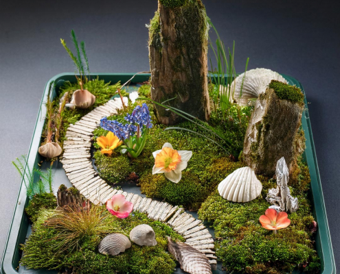 Fairy garden on a metal tray with seashells, feature rocks, flower heads and stick path.