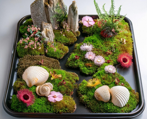 Fairy garden on a metal tray with seashells, feature rocks, fern and flower heads on mossy islands.